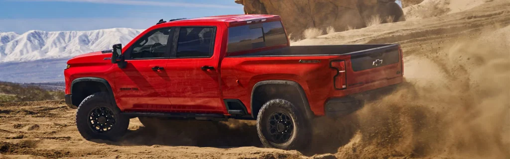 Chevrolet Silverado 3500 HD pickup truck driving off-road on a dirt terrain with dust kicking up and mountains in the background.